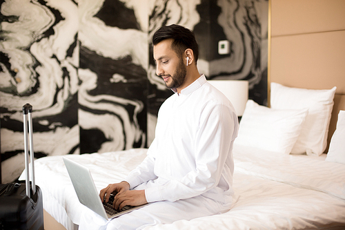 Young busy man with earphones and laptop sitting on bed and typing in hotel room or at home