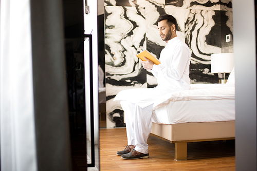 Contemporary man in white clothes sitting on his bed in hotel room or at home and reading business literature