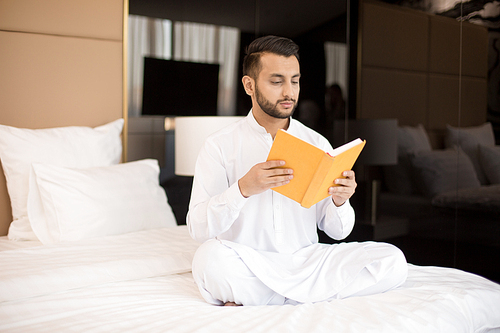 Cross-legged young man in white clothes sitting on bed and reading book at leisure