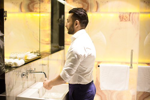 Young businessman standing by sink in front of mirror and washing hands in the morning