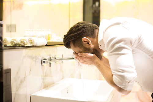 Young businessman washing his face over sink in hotel bathroom during his travel in the morning
