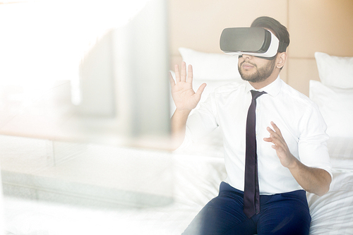 Modern businessman in formalwear sitting on bed and watching conference or presentation in virtual reality headset