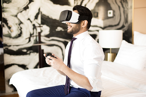 Young businessman in vr headset holding joystick while playing virtual games in hotel room