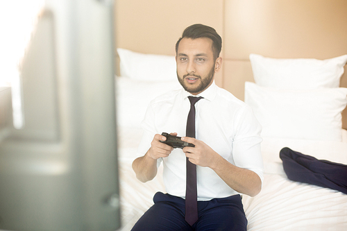 Businessman in formalwear sitting on bed in front of monitor and gaming in hotel room at break