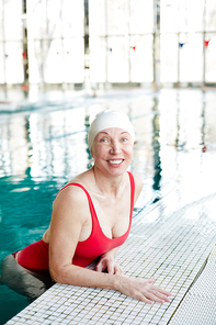 Cheerful mature female in red swimsuit and white rubber cap spending leisure in swimming pool
