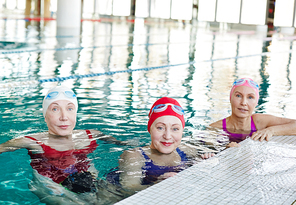 Three healthy mature women in swimwear sitting in water of swimming-pool before training