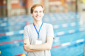 Happy successful trainer of swimming with badge hanging on his neck and crossed arms on chest looking at camaera on background of swimming-pool