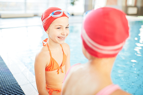 Happy girl in swimsuit, swim-cap and goggles looking at her friend while talking to her by swimming-pool