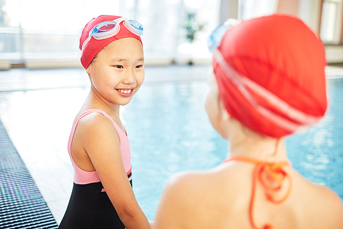 Cute Asian girl in swim-cap and goggles talking to her friend in swimming-pool