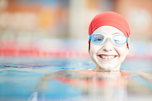 Happy young swimmer in goggles and swim-cap looking out of water of swimming-pool