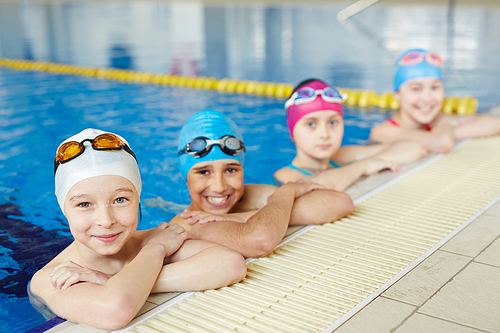 Group of joyful boys and girls smiling to camera at border of swimming pool during practice