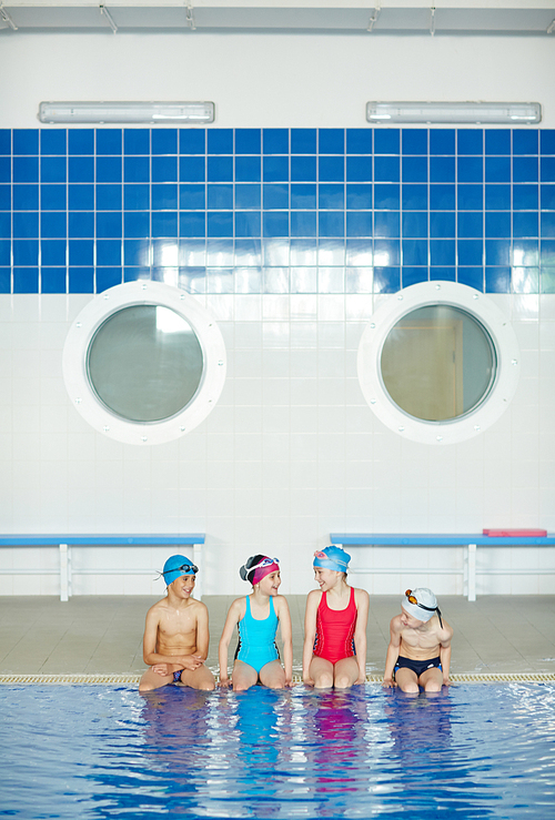 Portrait of cheerful school kids sitting at border of pool talking and laughing while waiting for swimming lesson