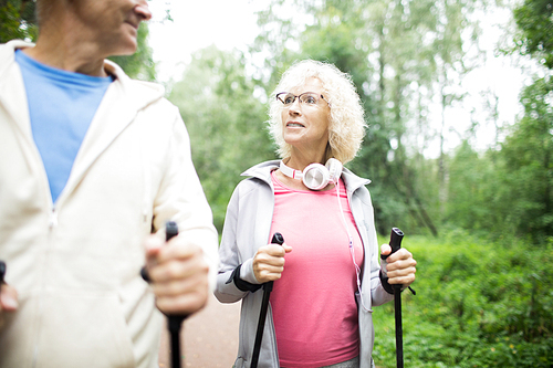 Active senior blonde woman in eyeglasses and sportswear trekking with her husband on summer day