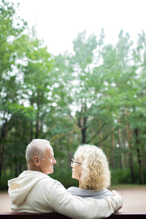 Back view of mature affectionate man and woman sitting on bench in park on summer day
