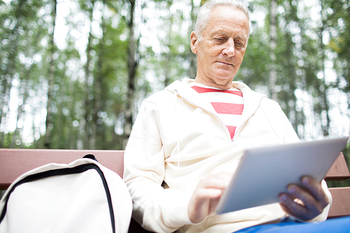 Active senior man with touchpad networking while sitting on bench in park and having rest