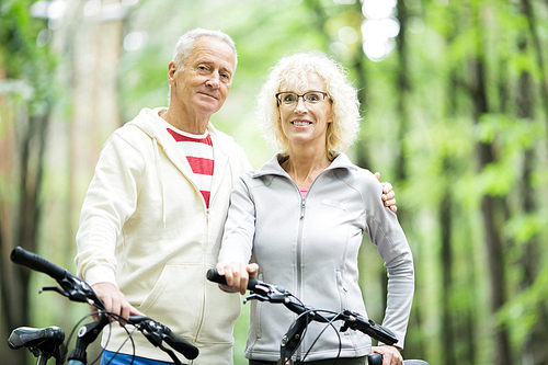 Affectionate and sporty senior couple looking at you while chilling out in park or forest with bicycles