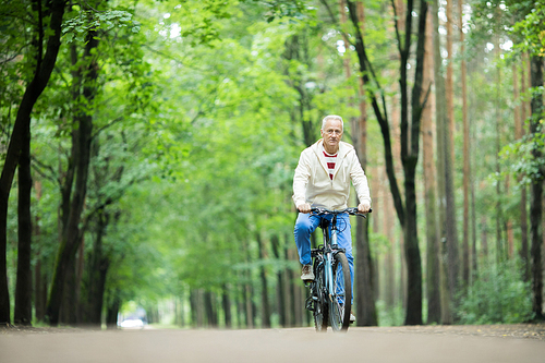 Retired active man riding bicycle along forest road between greeen trees on summer day