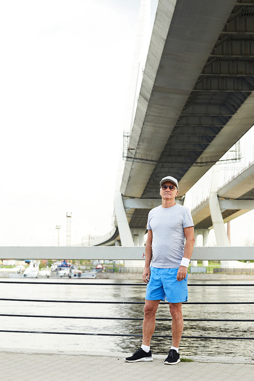 Senior sportsman in activewear and sunglasses standing by waterside under bridge in urban environment