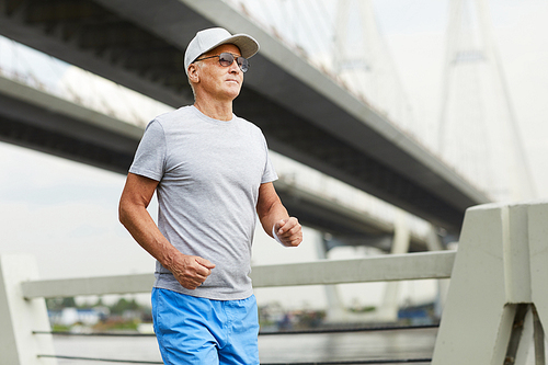 Aged man in sunglasses and sportswear jogging along riverside in the morning