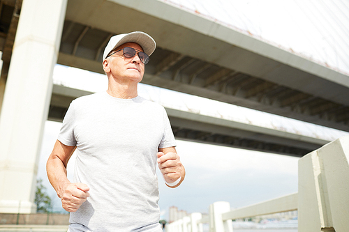 Mature man in sunglasses, grey t-shirt and baseball cap jogging in urban environment