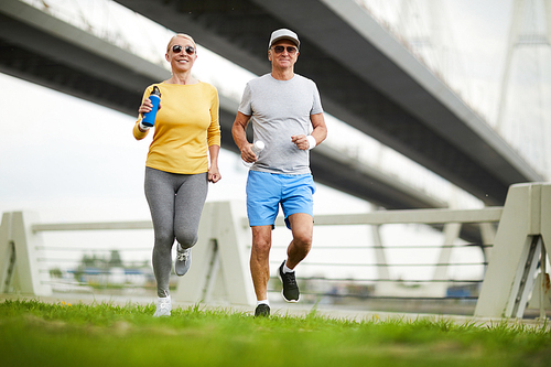 Happy and active aged couple in sportswear running on green lawn in urban environment