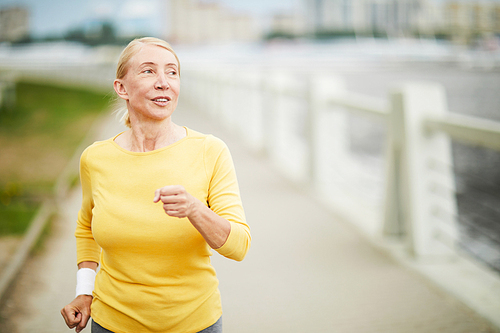 Aged blond female in activewear practicing morning jogging along riverside