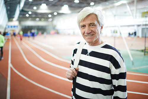 Happy senior man in casual striped pullover standing on stadium on background of tracks