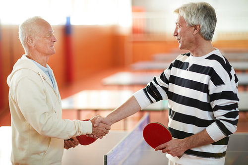 Two successful mature ping pong professionals in activewear shaking hands in tennis hall after game