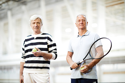 Two active senior men in casualwear holding tennis rackets and  on stadium