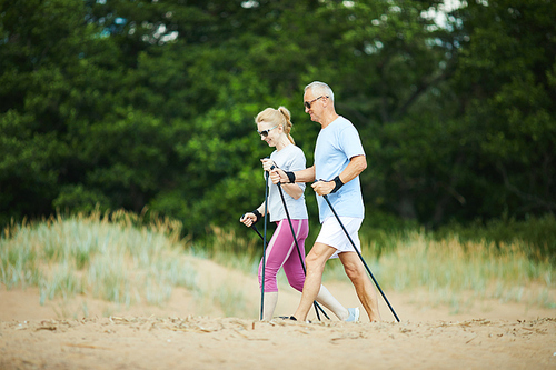 Modern senior couple in sunglasses and activewear trekking on sandy beach with green trees on background