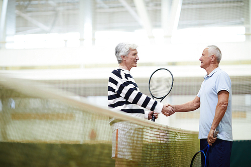 Two mature active tennis players with rackets handshaking over net after or before game on the court