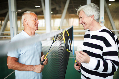 Two friendly senior badminton players in activewear with rackets standing by net and interacting