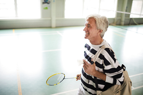 Happy aged active man with badminton racket and backpack leaving sports center after game