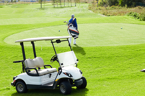 Golf car standing on green field and rear view of aged man with golf clubs walking to play