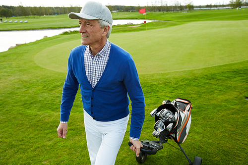 Active mature man in casualwear pulling sportive bag with golf clubs while walking down green lawn
