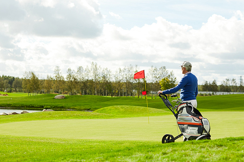 Aged golf player with sportive bag standing on large green play field on summer weekend