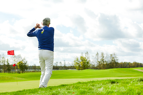 Rear view of active aged man in casualwear with golf club before hitting ball during play