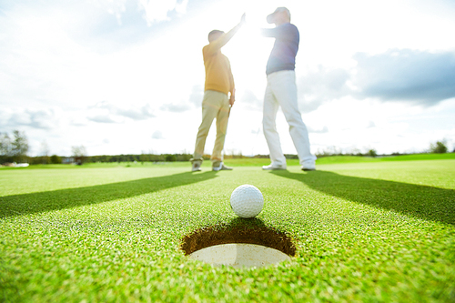 Golf ball lying on green play field close to hole with two successful players making high five on background