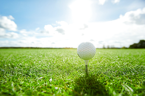 White golf ball on tee on vast green play field with cloudy sky and sunshine above