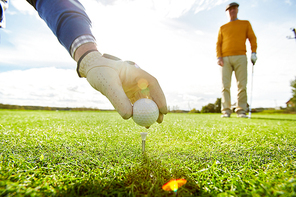 One of golf players putting ball on tee while the other one standing on background