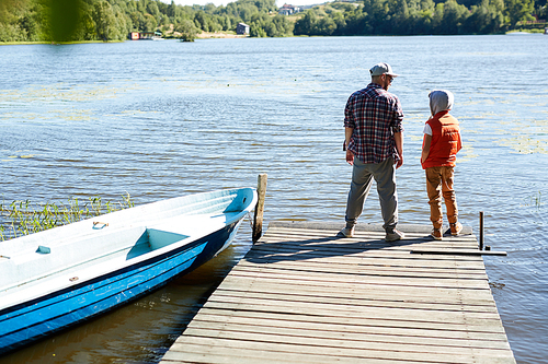 Rear view of dad and son standing on wooden pontoon in front of waterside with boat near by