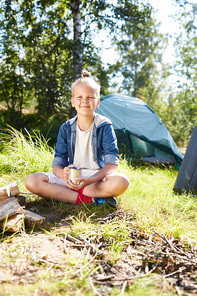 Little backpacker with cup of tea sitting on grass by campfire on background of his tent