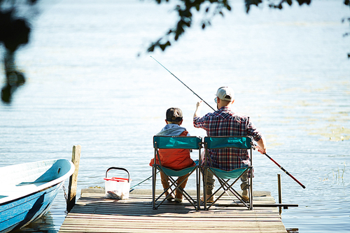 Rear view of man and his son sitting in front of water and fishing together at summer weekend