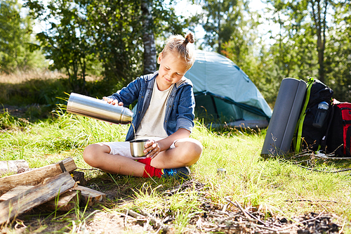 Youthful boy pouring tea in mug while enjoying summer day by campfire on background of his tent