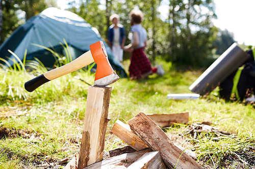 Axe in piece of firewood prepared for campfire with scouts by tent on background