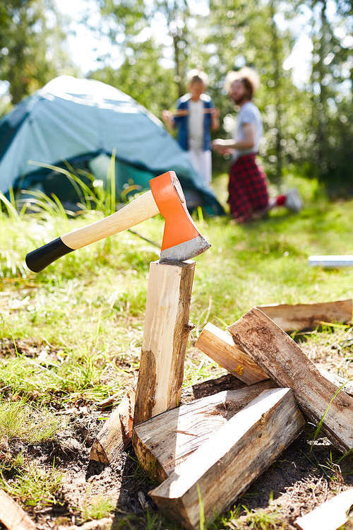 Axe in one of wooden pieces ready for making bonfire and tourists by tent on background
