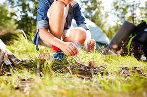 Boy with matches making fire and burning firewood on grass on summer day during trip
