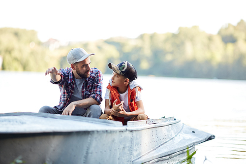 Young man pointing forward while sitting in boat with his son and explaining something