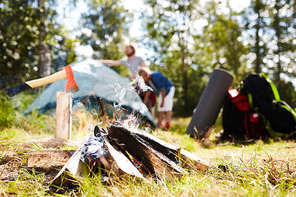 Smoke and fire of burning firewood on grass with tent, backpacks and tourists on background