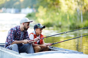Father and son in casualwear sitting in boat with rods and fishing together on weekend